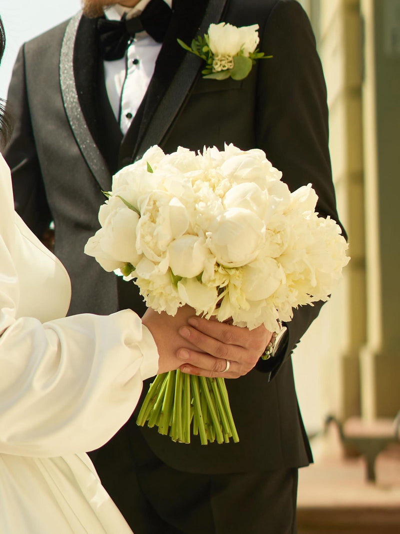 Brautstrauss aus weissen Pfingstrosen, gehalten von der Braut und dem Bräutigam in eleganter Hochzeitskleidung.