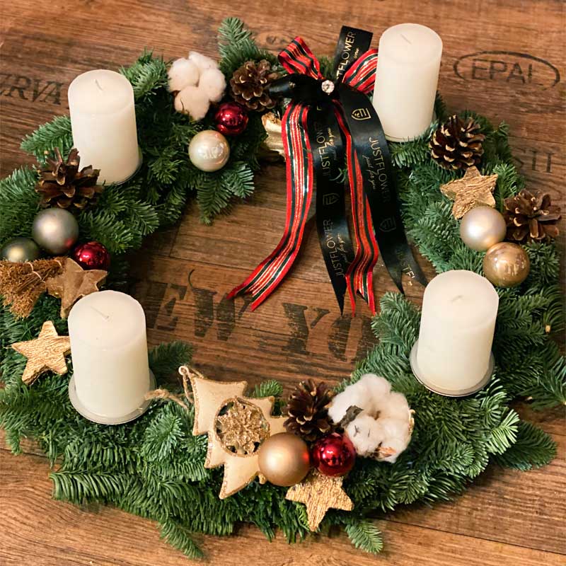 Adventskranz aus frischen Tannenzweigen, dekoriert mit weißen Kerzen, roten und goldenen Kugeln, Sternen aus Gold, Tannenzapfen und Baumwolle. Ein festliches JustFlower-Schleifenband vervollständigt das elegante Design.